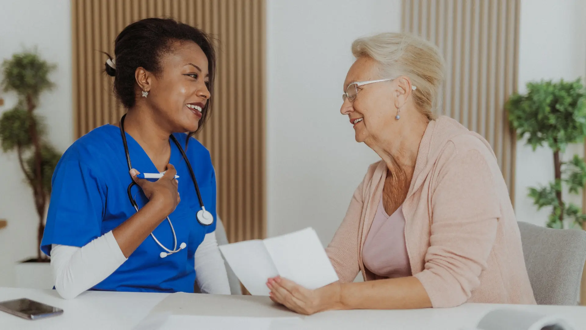 A healthcare professional in blue scrubs talking with an older woman holding notes during a doctor visit.