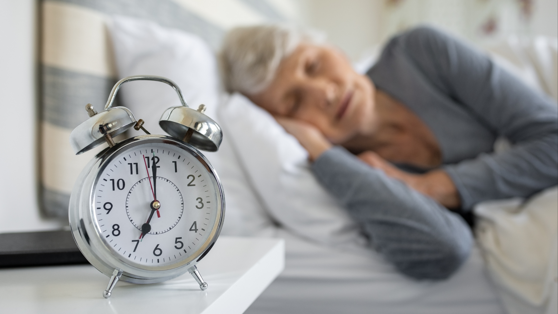 An alarm clock beside an older adult resting in bed.
