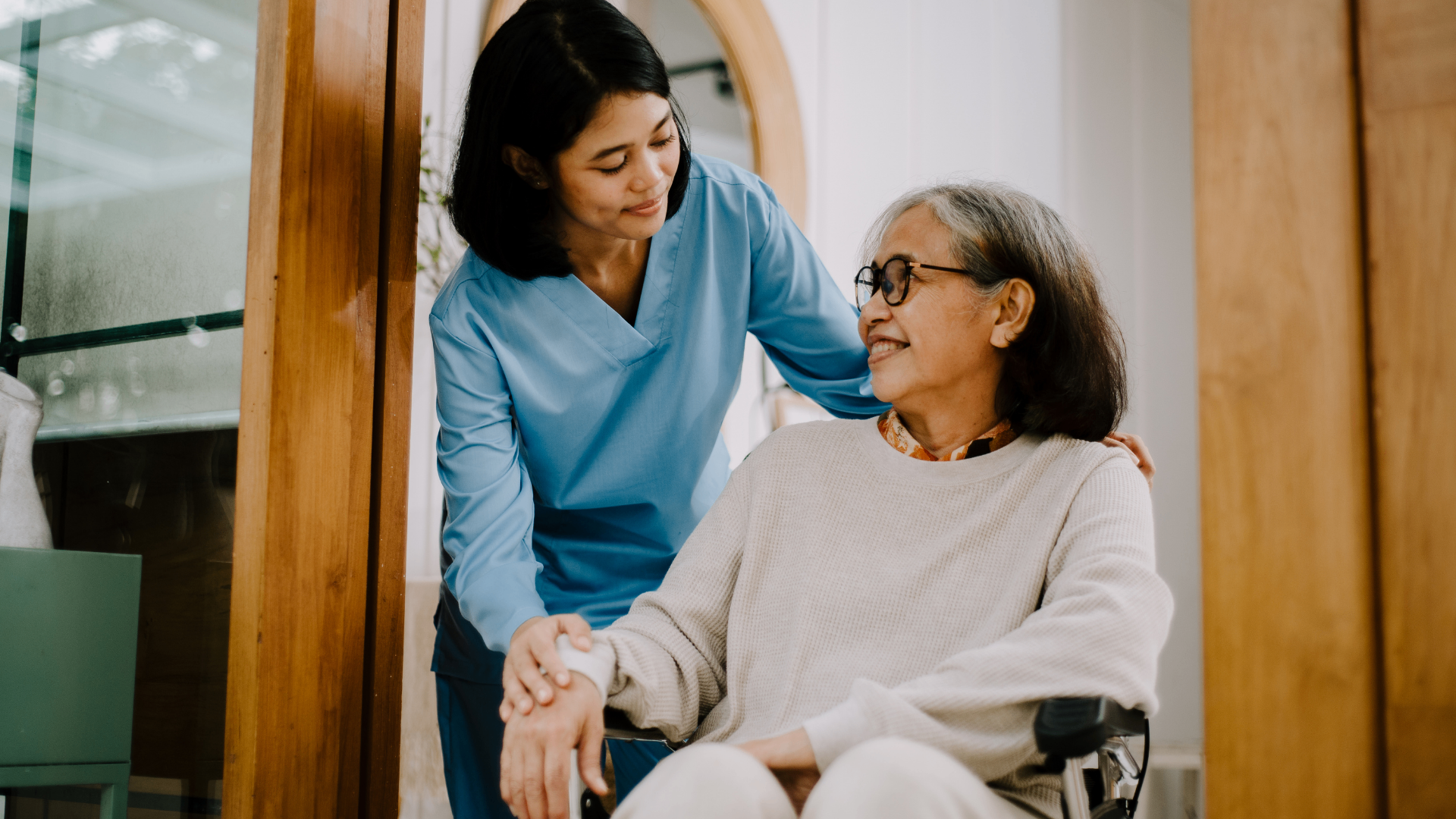 A Troy Medicare Care Manager in blue scrubs smiles while gently supporting an older adult member seated in a wheelchair at home.