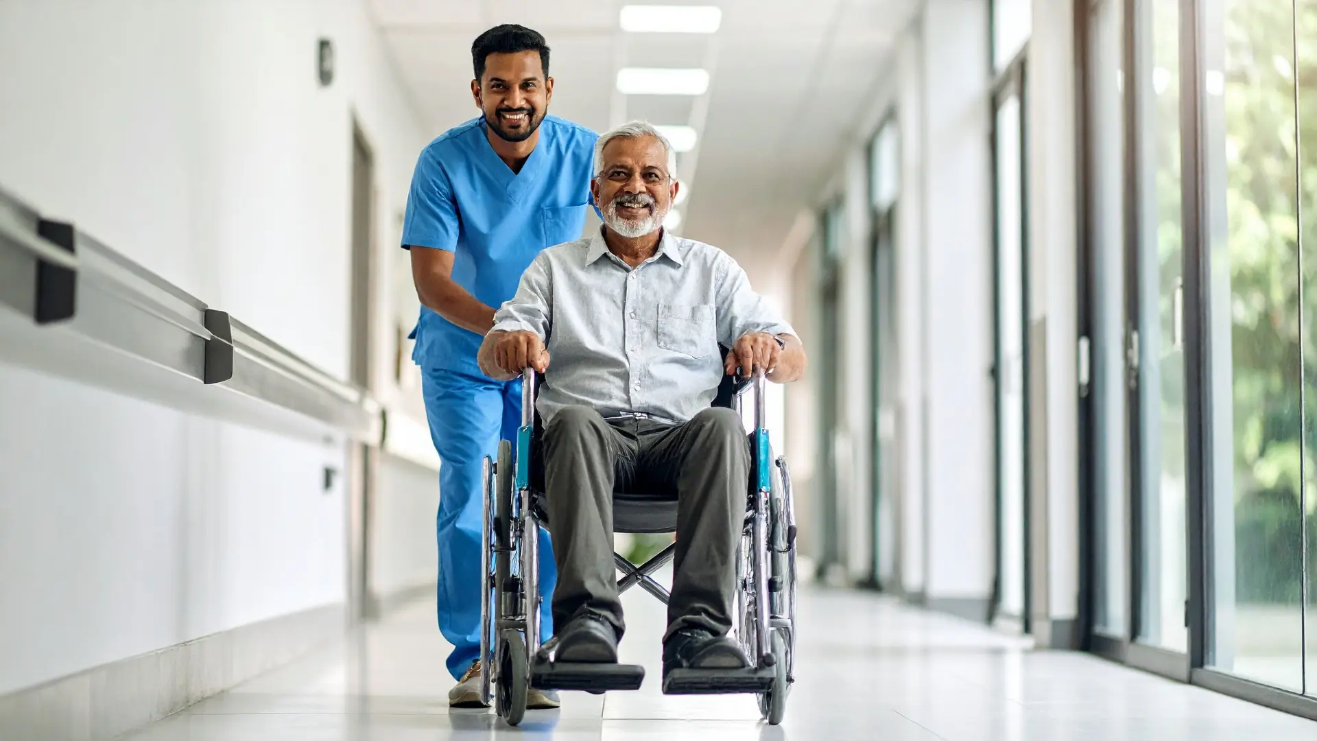 A healthcare worker in blue scrubs smiling while pushing an older man in a wheelchair down a bright hospital hallway.