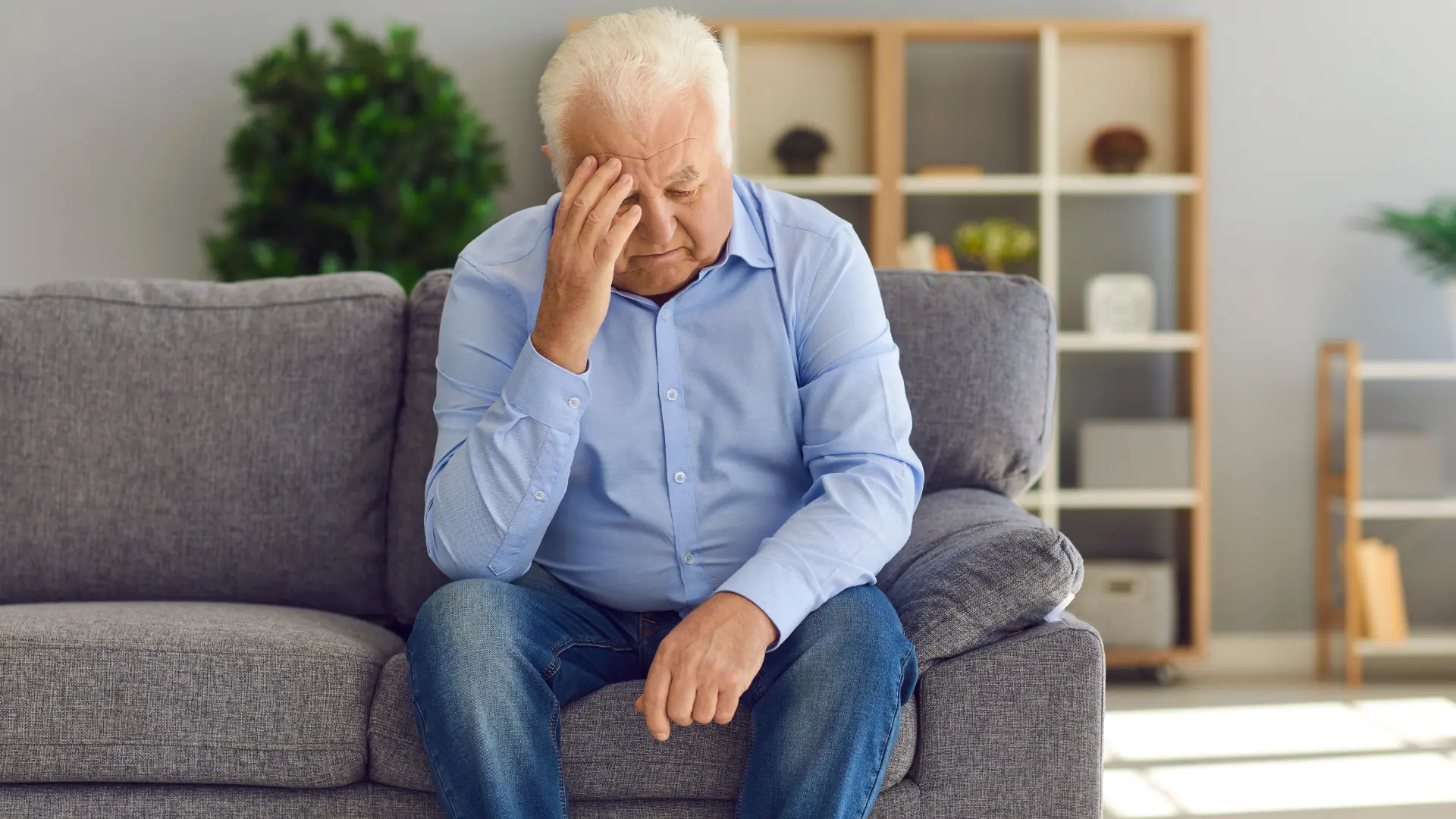 An older man sitting on a grey couch at home, resting his hand against his head.