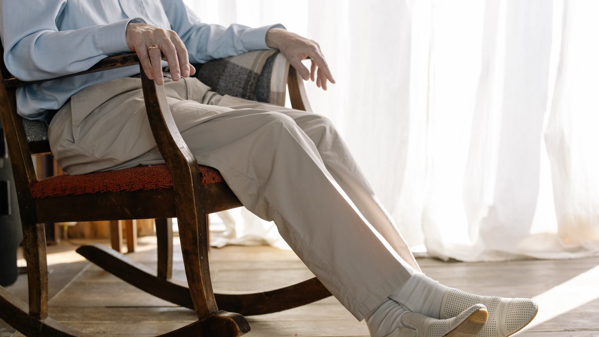 An older adult resting comfortably in a wooden rocking chair beside a sunlit window with sheer curtains.