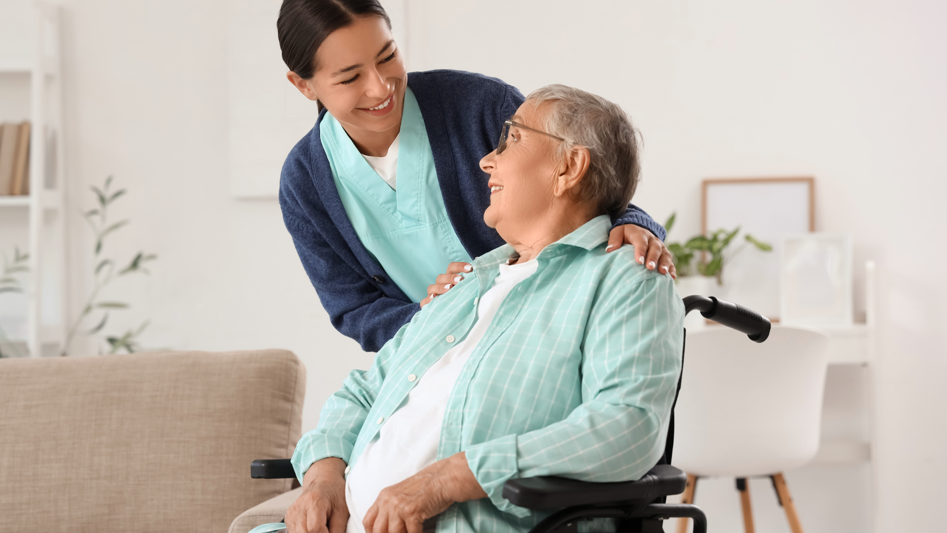 A nurse practitioner in teal scrubs warmly assisting an older woman in a wheelchair in a bright home setting.